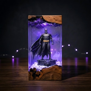 Puede incluir: Una pieza decorativa rectangular de resina y madera con una figura de Batman sobre una base de g&aacute;rgola. El interior de resina transparente est&aacute; iluminado con un brillo p&uacute;rpura, creando un efecto estrellado tras la figura.