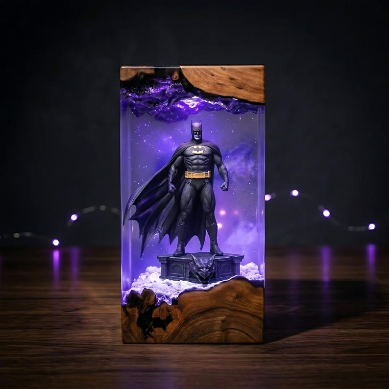 Puede incluir: Una pieza decorativa rectangular de resina y madera con una figura de Batman sobre una base de g&aacute;rgola. El interior de resina transparente est&aacute; iluminado con un brillo p&uacute;rpura, creando un efecto estrellado tras la figura.
