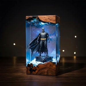 May include: A rectangular lamp featuring a Batman figurine standing on a gargoyle base, encased in clear blue resin with embedded white clouds and a starry night effect, framed by natural wood edges at the top and bottom of the structure.