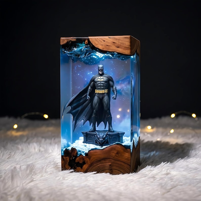 Puede incluir: Una l&aacute;mpara rectangular con una figura de Batman incrustada en resina azul transparente, con bordes de madera natural arriba y abajo. La figura se alza sobre un pedestal oscuro, iluminada desde el interior con un fondo estrellado y nebuloso.