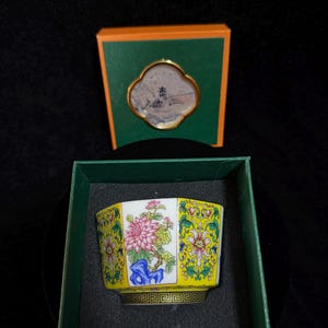 May include: A hexagonal porcelain bowl featuring a vibrant yellow background with intricate floral patterns in pink, green, and blue, set inside a dark green box. A matching orange and green box lid with a circular landscape illustration is placed behind.