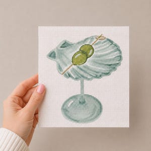 May include: A cross-stitch embroidery piece on white fabric featuring a light blue cocktail glass shaped like a seashell. The glass contains two green olives on a wooden pick, rendered with detailed stitching to create a textured appearance.