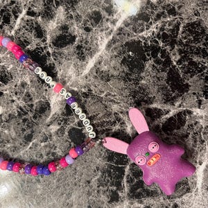 Könnte beinhalten: Ein Perlenband in den Farben Violett, Pink und Helllila mit weißen Buchstabenperlen, die JUDGEE AS FUGG buchstabieren. Am Ende befindet sich ein violetter Anhänger in Hasenform mit rosa Innenohren, Knopfaugen und einem orangefarbenen Mund.