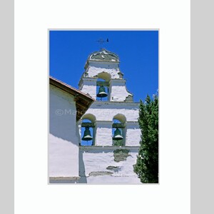 May include: A white bell tower with three arched openings, each housing a bell. The tower has a cross on top and is set against a bright blue sky. A portion of a building is visible on the left.