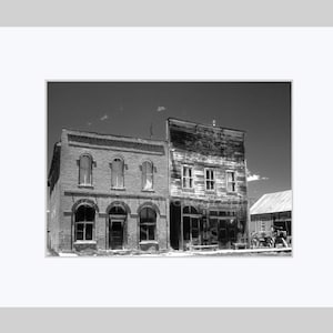 May include: Black and white photograph of two weathered buildings in a rural setting. The building on the left is brick with arched windows and doorways. The building on the right is wooden and appears to be in disrepair.