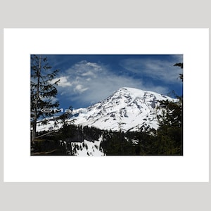 May include: A framed landscape photograph of a snow-covered mountain under a blue sky with wispy clouds. The mountain is surrounded by evergreen trees. The photograph is framed in white.