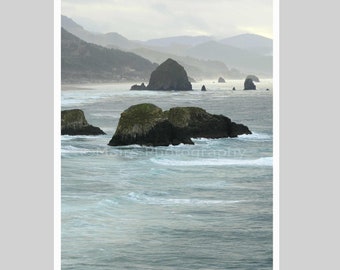 May include: A coastal landscape featuring dark, moss-covered rock formations rising from the ocean waves. In the distance, a large monolith stands near a sandy beach, with hazy, layered mountains under a soft, overcast sky. The text Mairs Photography is visible.