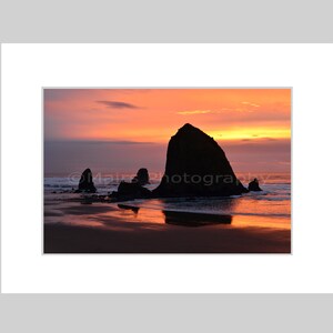 May include: A scenic photograph of a coastal landscape at sunset. Silhouetted rock formations stand in the ocean, reflecting the vibrant orange and pink hues of the sky and water. The image is framed with a white mat.