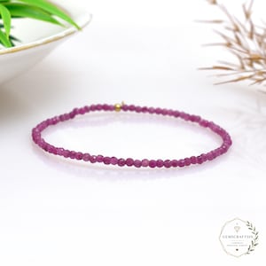 May include: A delicate bracelet composed of small, faceted, deep pink ruby beads arranged in a continuous circular strand. A single, tiny, polished gold-tone metal bead is positioned near the clasp area, providing a subtle metallic accent.