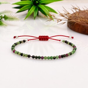 Pode incluir: Uma pulseira delicada com pequenas contas de pedras preciosas facetadas em verde e magenta dispostas num padrão repetitivo. A pulseira inclui um cordão vermelho com um nó deslizante ajustável e pequenas contas metálicas prateadas.