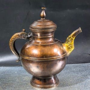 Könnte beinhalten: Eine bronzefarbene Teekanne aus Metall mit rundem Körper, Standfuß und Deckel mit Knauf. Der Griff ist als strukturierter Drache geformt, während der Ausguss mit einem Drachenkopf-Design in einem helleren Metallton verziert ist.
