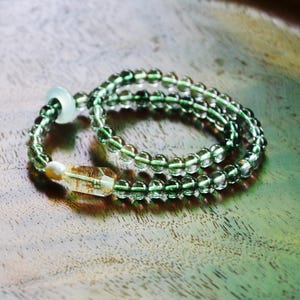 May include: A wrap bracelet composed of translucent green spherical beads, featuring a single cylindrical clear quartz bead with golden inclusions, a small white pearl, and a pale green circular accent bead strung on a flexible cord.
