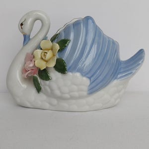 May include: A white ceramic swan figurine featuring a blue-tinted wing section with molded feather details. The swan is decorated with a cluster of small, sculpted yellow and pink roses with green leaves positioned on the side of its neck.