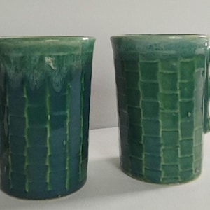 May include: Two ceramic mugs with a glossy green glaze and a textured basket-weave pattern on the exterior. Each mug features a distinctive handle design consisting of two stacked circular loops, providing a unique grip for the user.