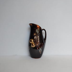 May include: A dark brown ceramic pitcher with a glossy finish, featuring a raised relief design of golden-toned leaves and round fruit on the front. The pitcher has a curved handle, a tapered spout, and a smooth, rounded body shape.