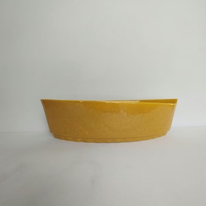 May include: A mustard yellow ceramic bowl with a speckled texture and a wide, shallow, elongated oval shape. The base of the bowl features a decorative scalloped edge detail that runs along the bottom perimeter of the vessel.