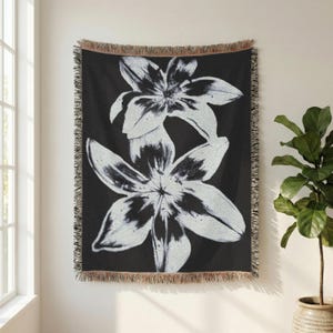 May include: A rectangular woven throw blanket featuring a high-contrast black and white graphic of two large lily flowers. The blanket has a textured, fringed edge along the top and bottom, with a dark background and light floral details.