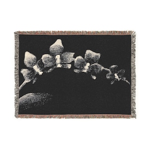 May include: A rectangular woven throw blanket featuring a black background with a large, off-white orchid flower branch design. The blanket has a textured, fringed edge with multi-colored threads visible along the top and bottom borders.