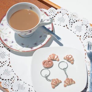 May include: A white porcelain teacup filled with light brown tea sits on a matching saucer with floral patterns. Next to it, a white square plate holds two croissant keychains, one loose croissant charm, one pretzel charm, and a silver fork.