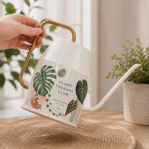 May include: A white metal watering can with a gold-toned handle and a long, slender spout. The exterior features a botanical print of green monstera leaves, a string of hearts vine, and the text "PLANT THERAPY CLUB BREATHE GREEN LIVE SLOW."