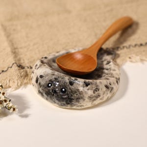 Könnte beinhalten: Eine Keramik-Löffelablage in Form einer gefleckten Robbe mit glänzender, weiß-grau gesprenkelter Oberfläche, verziert mit aufgemalten schwarzen Augen und Schnurrhaaren. Ein glatter, hellbrauner Holzlöffel liegt in der Vertiefung der Schale.