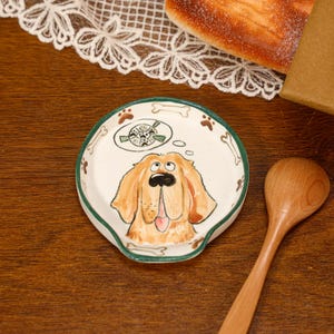 May include: A round ceramic spoon rest with a green rim, featuring a hand-painted golden-brown dog with a black nose and tongue, looking up at a thought bubble containing a ball with paw prints, surrounded by brown paw prints and white bones.