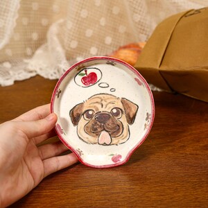 Puede incluir: Un reposacucharas redondo de cerámica con superficie color crema moteada que presenta una ilustración pintada a mano de un perro pug con la lengua fuera. Sobre el pug hay una burbuja de pensamiento con una manzana roja. El borde es rojo oscuro.