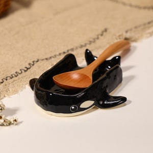 May include: A glossy black and white ceramic spoon rest shaped like an orca whale, featuring a smooth finish and a small white eye detail. A light brown wooden spoon with a rounded bowl rests inside the curved body of the ceramic whale figure.
