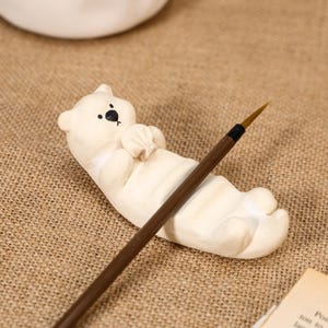 Può includere: Una statuetta di orso in ceramica color crema a forma di poggia pennello, con un piccolo naso e occhi neri, su cui poggia un pennello con manico marrone. L'orso è raffigurato in posizione sdraiata con le zampe tenute vicino al viso.