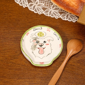 Puede incluir: Un reposacucharas redondo de cerámica con una ilustración pintada a mano de un perro blanco con una oreja gris, lengua rosa y una burbuja de pensamiento con una pelota de tenis verde. El borde verde tiene pequeños huesos blancos y huellas rosas.