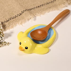May include: A yellow ceramic spoon rest shaped like a duck with a light blue center, featuring a smooth glossy finish and black eyes. A small, light brown wooden spoon rests inside the duck-shaped dish, which has a rounded body and wings.