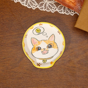 Puede incluir: Un reposacucharas redondo de cerámica con borde amarillo, que presenta una ilustración dibujada a mano de un gato blanco y naranja con collar y una burbuja de pensamiento que contiene una campana. La superficie tiene huellas de patas naranjas.