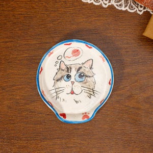 Puede incluir: Un reposacucharas redondo de cerámica con base color crema y borde azul. El centro presenta una ilustración pintada a mano de un gato esponjoso con grandes ojos azules y una burbuja de pensamiento que contiene un ovillo de lana rojo. Corazones y huellas rojas decoran el borde.