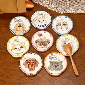 Op de afbeelding: Een verzameling van acht ronde keramische lepelhouders, elk voorzien van een handgeschilderde illustratie van een ander dierengezicht, waaronder honden, katten en een konijn, met gedachtewolkjes die diverse items zoals botten en ballen tonen.