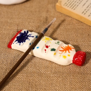 Op de afbeelding: Een witte keramische verftube-houder met een rode dop en rood uiteinde, versierd met kleurrijke verfspatten in blauw, geel, rood, groen en oranje. Een dunne bruine verfkwast met een doorzichtige plastic punt rust op de keramische houder.
