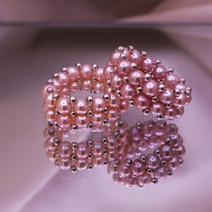 May include: A ring constructed from multiple rows of small, round, light pink pearls interspersed with tiny, reflective silver-toned metallic beads. The design features a dense, textured arrangement of pearls that creates a wide, circular band.
