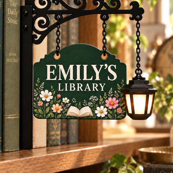 May include: A dark green rectangular sign with the white text EMILY'S LIBRARY, featuring a floral border and an open book illustration. The sign hangs from a black metal scrollwork bracket with a small attached lantern emitting a warm light.