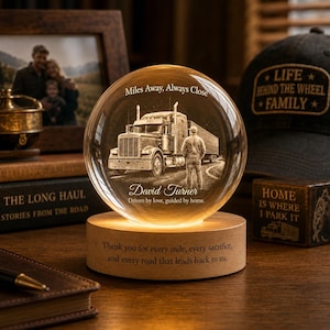 May include: A spherical glass paperweight on a round wooden base. The glass features an etched image of a semi-truck and a person standing nearby, with the text "Miles Away, Always Close", "David Turner", and "Driven by love, guided by home."