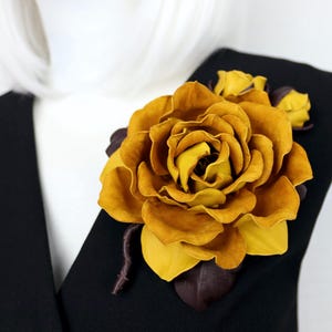 May include: A large, handcrafted yellow rose brooch with detailed petals and smaller buds. The brooch is made of leather and features dark brown leaves. It is pinned to a black garment, creating a striking contrast.