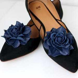 May include: Black flats with navy blue leather flower detailing. The shoes have a pointed toe and a suede-like finish. The floral embellishments add a decorative touch to the otherwise simple design, suitable for various occasions.