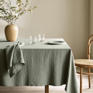 May include: A sage green linen tablecloth draped over a rectangular table, featuring a matching folded napkin on the corner. A beige ceramic vase with flowering branches, two clear glass tumblers, and a small grey plate sit on the tabletop.