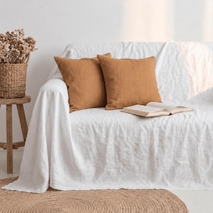 May include: Two square, solid-coloured, ochre-brown throw cushions are placed on a white sofa covered with a white fabric throw. An open book rests on the sofa, and a woven basket filled with dried flowers sits on a wooden stool nearby.