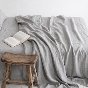 May include: A light grey linen bed sheet covering a mattress with an open book resting on top. Beside the bed stands a small, weathered wooden stool with four legs and a thick, rectangular seat featuring a visible wood grain texture.