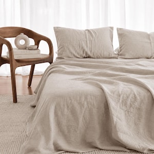 May include: A bed set featuring a light beige, textured linen duvet cover and two matching pillowcases. Beside the bed, a dark brown wooden chair holds two stacked books, a small ceramic bowl, and a round, off-white decorative vase.