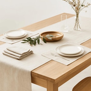 Peut inclure: Un chemin de table en lin beige clair et des sets de table assortis sur une table en bois. La disposition comprend des assiettes en céramique blanche, une pile de serviettes beiges pliées, un bol en bois, un vase en verre et un verre.