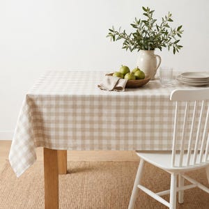 May include: A rectangular table covered with a beige and white gingham tablecloth. On the table sits a ceramic pitcher filled with green leafy branches, a wooden bowl containing green pears, a folded beige napkin, a glass, and stacked plates.