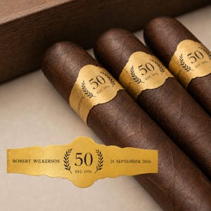 May include: Three dark brown cigars featuring gold paper bands. Each band displays the number 50, the text EST. 1976, and a laurel wreath design. A separate gold band shows the text ROBERT WILKERSON, 50, EST. 1976, and 21 SEPTEMBER 2026.