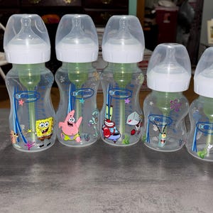 May include: Five Dr. Brown's baby bottles with clear plastic bodies, white screw-on caps, and translucent lids. Each bottle features internal green vent inserts and colorful graphics of SpongeBob SquarePants characters including Patrick Star and Mr. Krabs.