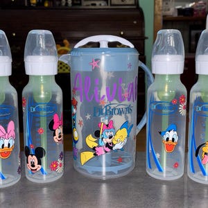 May include: Four clear plastic Dr. Brown's baby bottles with white caps and green internal vents, featuring colorful Disney character graphics, alongside a matching clear plastic pitcher with a blue handle and lid, also decorated with Disney characters.