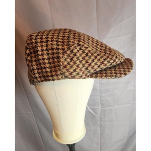 May include: A flat cap featuring a brown, tan, and dark red houndstooth pattern. The cap has a rounded crown and a short, stiff brim extending from the front. The textured fabric displays a classic woven appearance with a multi-colored check design.